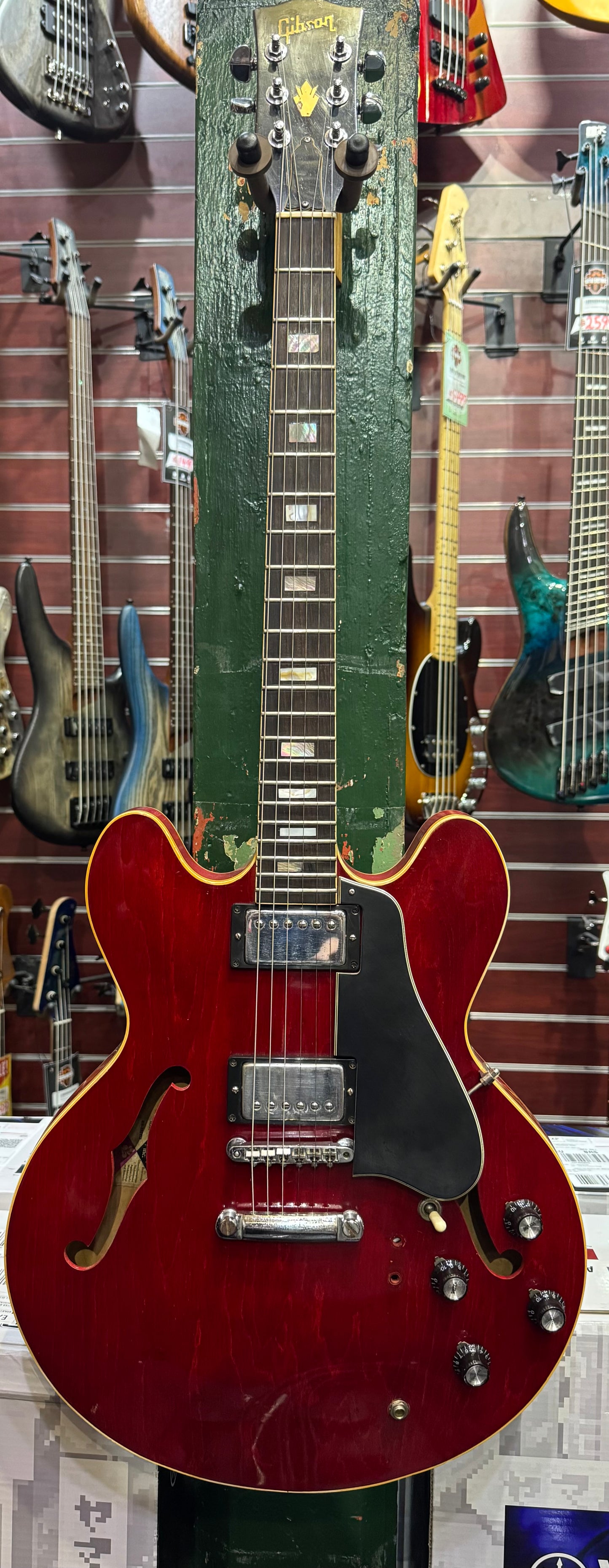Red electric guitar with a green pickguard on display against a wall of other guitars.